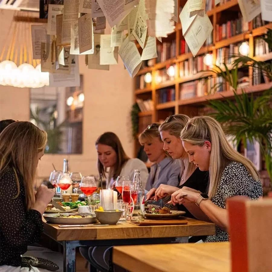 Guests eating together at a long table in The Bookshop, under hanging book pages and pendant lights