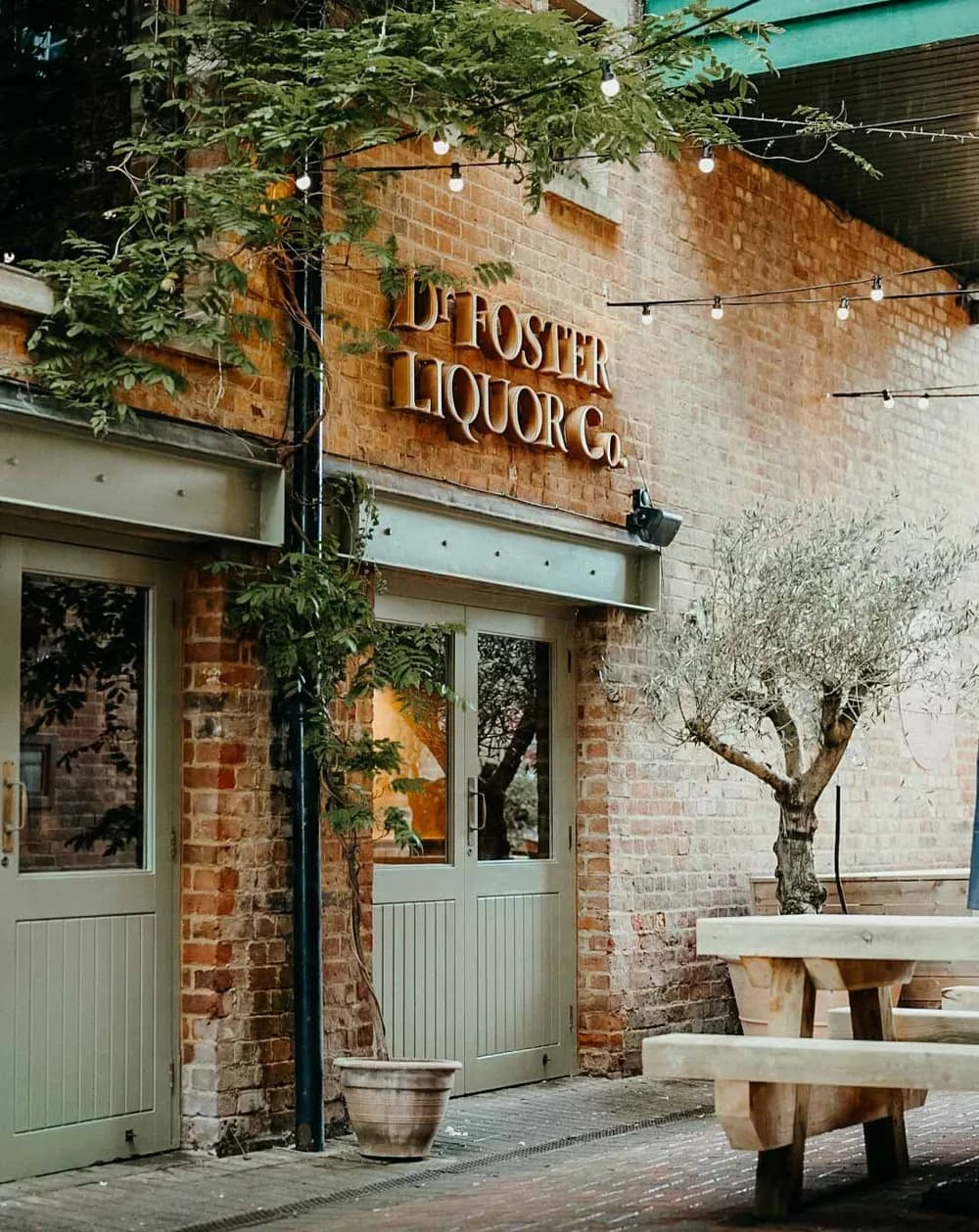 The brick frontage of Dr Foster, with an olive tree and a picnic bench outside