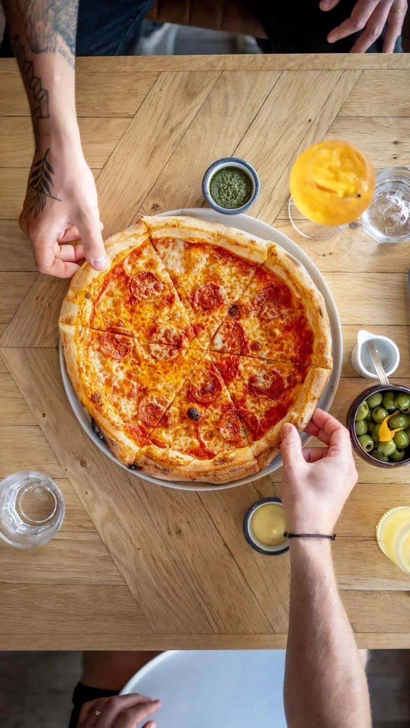 A pizza on a wooden table seen from above, with two people reaching in