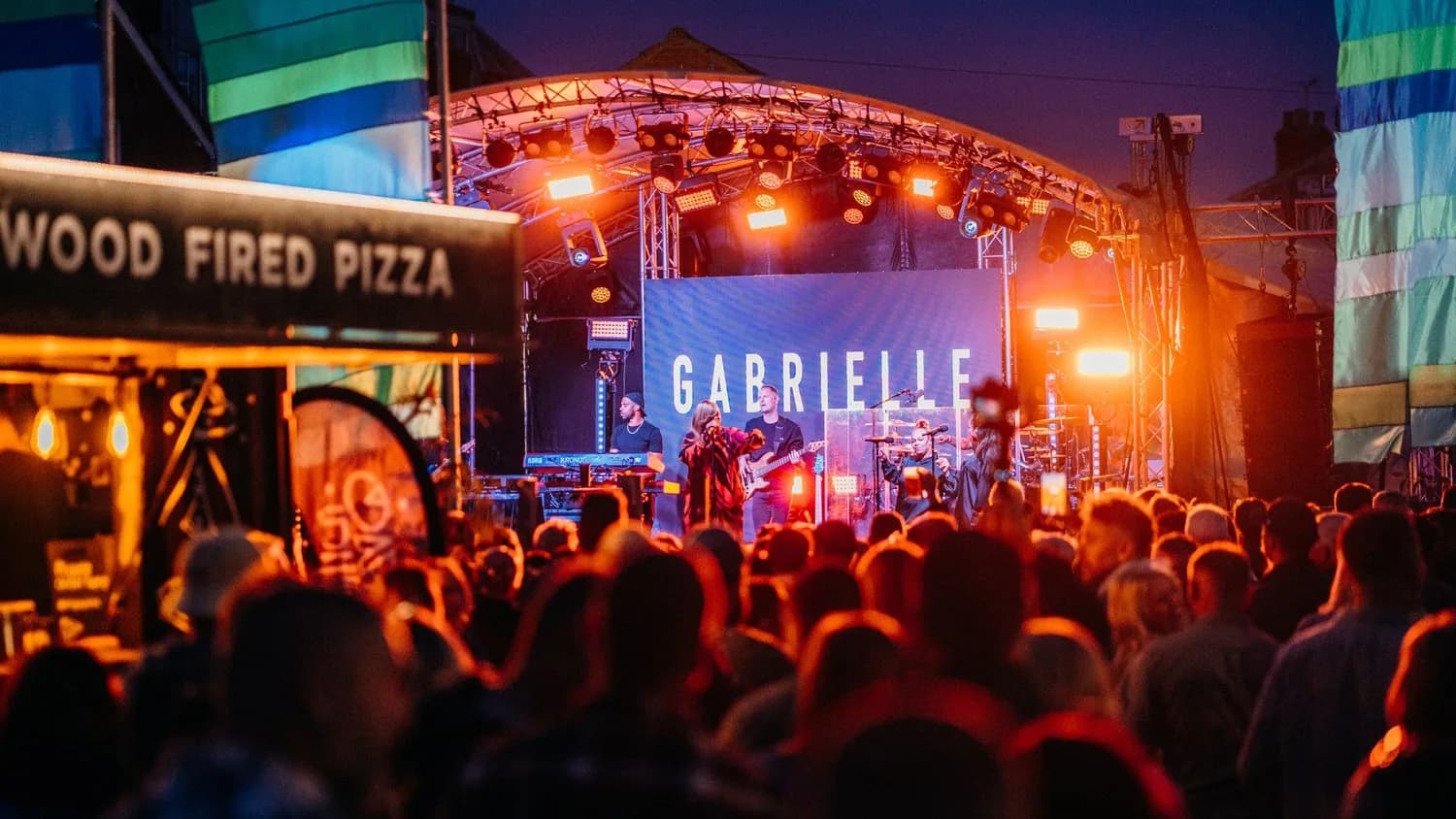 A crowd in front of an outdoor stage at a live music night, beside a wood-fired pizza counter