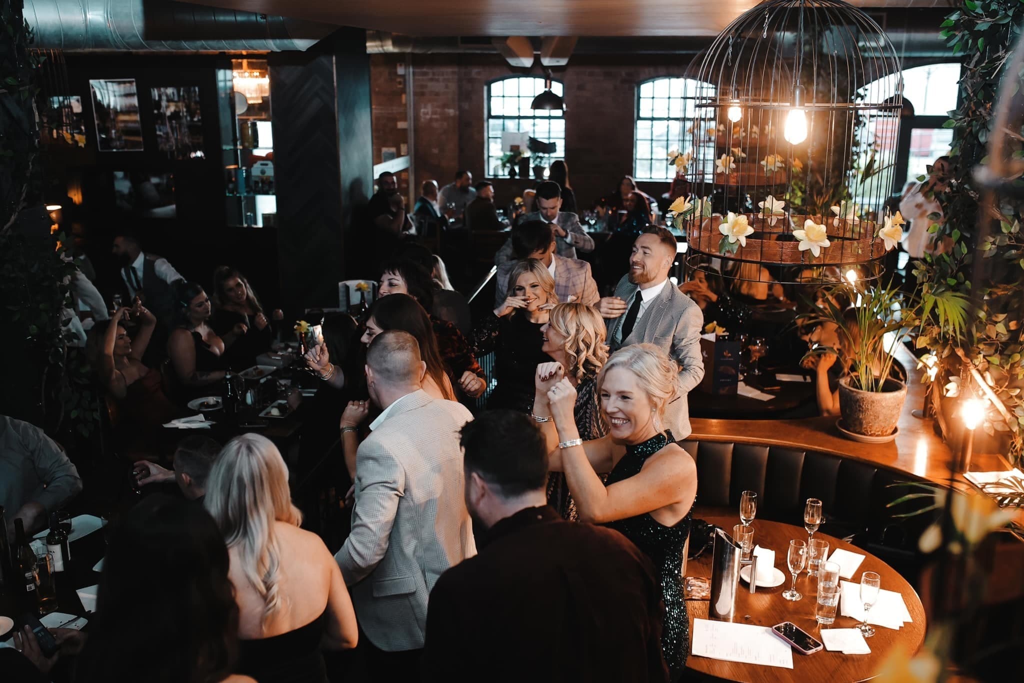 A packed dining room with guests dancing and celebrating between the tables, under a giant birdcage light